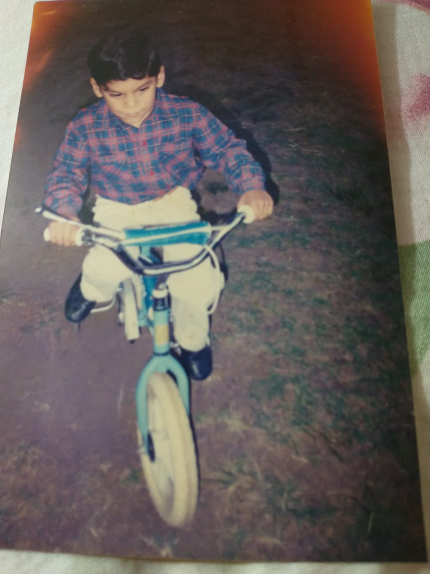 Yo, a los 7 años, en mi primer bicicleta con rueditas. Tengo el pelo corto, una camisa a cuadritos y jeans beige