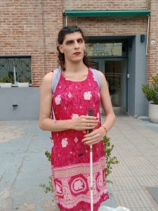 Katherine de pie frente a la entrada de un edificio de ladrillo, mirando hacia adelante con expresión serena. Lleva un vestido rojo estampado, maquillaje con labios rojos y el cabello oscuro recogido hacia atrás. Sostiene un bastón blanco mientras lleva una mochila en los hombros, destacando el rojo del vestido frente al fondo del edificio.
